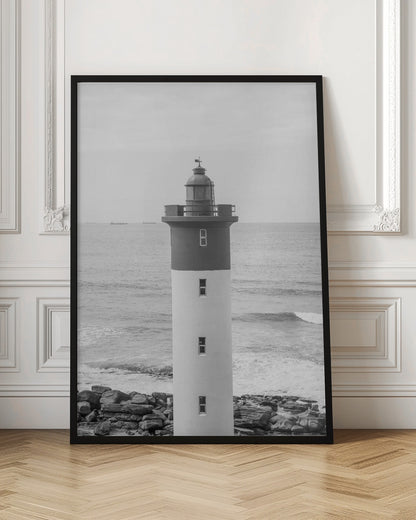 A framed, black and white photograph of a tall, cylindrical lighthouse standing on a rocky shore. The lighthouse is dark grey on its upper section and white on the lower, with the ocean and a grey sky in the background. Print