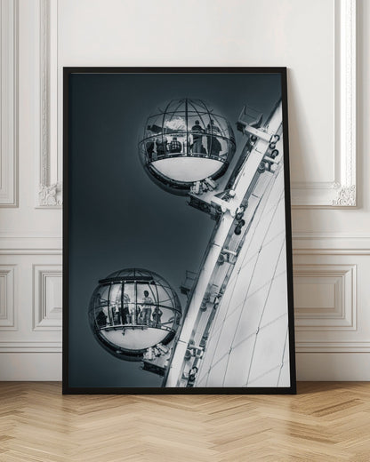 A dramatic low-angle, black and white photograph of two spherical glass pods of the London Eye Ferris wheel. Silhouettes of people are visible inside the pods against a dark, solid sky, with the massive white structure of the wheel on the right. Print
