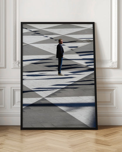 A man in a dark jacket and blue jeans stands alone in profile on a geometrically patterned plaza. The ground is a striking pattern of grey and white triangles, crossed by long, dark shadows cast by the bright sun, creating a high-contrast, graphic composition. Wall Art
