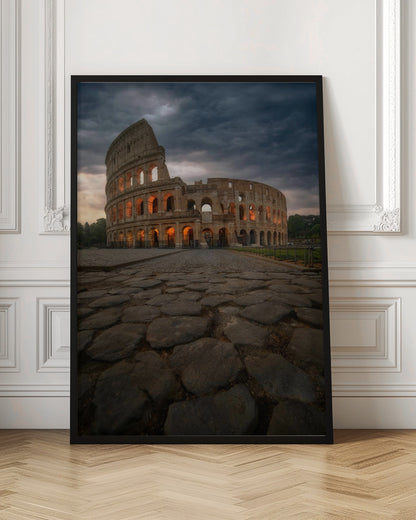 A low-angle view of the Roman Colosseum at dusk, with its arched windows glowing with warm orange light under a dramatic, stormy sky. An ancient cobblestone road leads towards the iconic amphitheater. Print