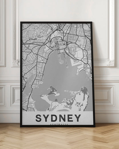 A modern, black and white framed art print of a stylized map of Sydney, Australia. The map features bold black lines for the street grid and major roads, with light grey for the landmass and a darker grey for the water of the harbor. The word 'SYDNEY' is printed in large, black, sans-serif font at the bottom of the poster. Poster