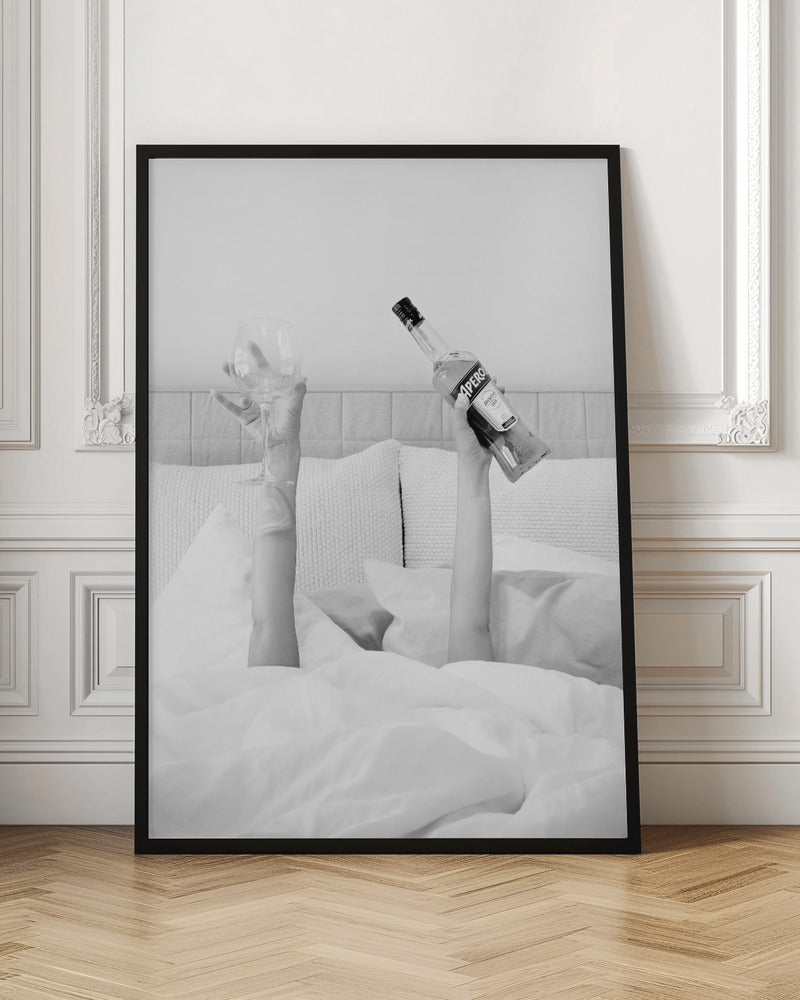 A black and white photograph of a person under white covers in bed, with only their arms visible. One hand holds up a bottle of Aperol, and the other holds up an empty wine glass. Decor