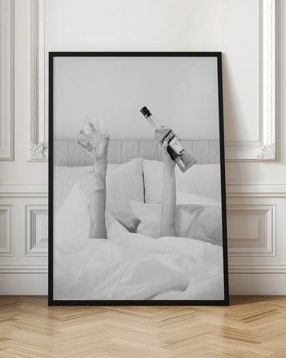 A black and white photograph of a person under white covers in bed, with only their arms visible. One hand holds up a bottle of Aperol, and the other holds up an empty wine glass. Decor