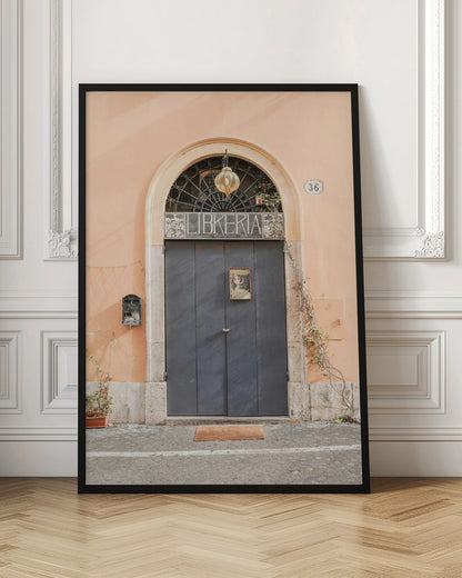 A charming European bookstore entrance featuring a large, dark grey arched door set against a peach-colored wall. Above the door, a sign reads 'LIBRERIA' below a decorative fanlight window and a hanging lantern. Decor