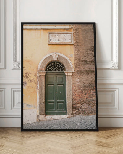 A vertical shot of a rustic European street scene featuring a dark green arched wooden door set into a stone frame. The building's facade is a mix of peeling yellow ochre plaster and exposed red brick. Above the door is a marble plaque that reads 'SALITA RI DEL GRILLO', and the ground is paved with grey cobblestones. Wall Art