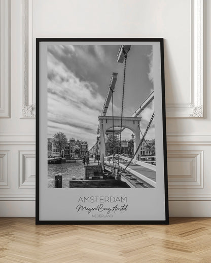 A black and white travel poster of Amsterdam, featuring a side view of the Magere Brug, a traditional wooden drawbridge, over the Amstel river. Classic canal houses line the waterway in the background under a dramatic, cloudy sky. The image is in a black frame and has text at the bottom that reads 'AMSTERDAM', 'Magere Brug, Amstel', and 'NEDERLAND'. Artwork