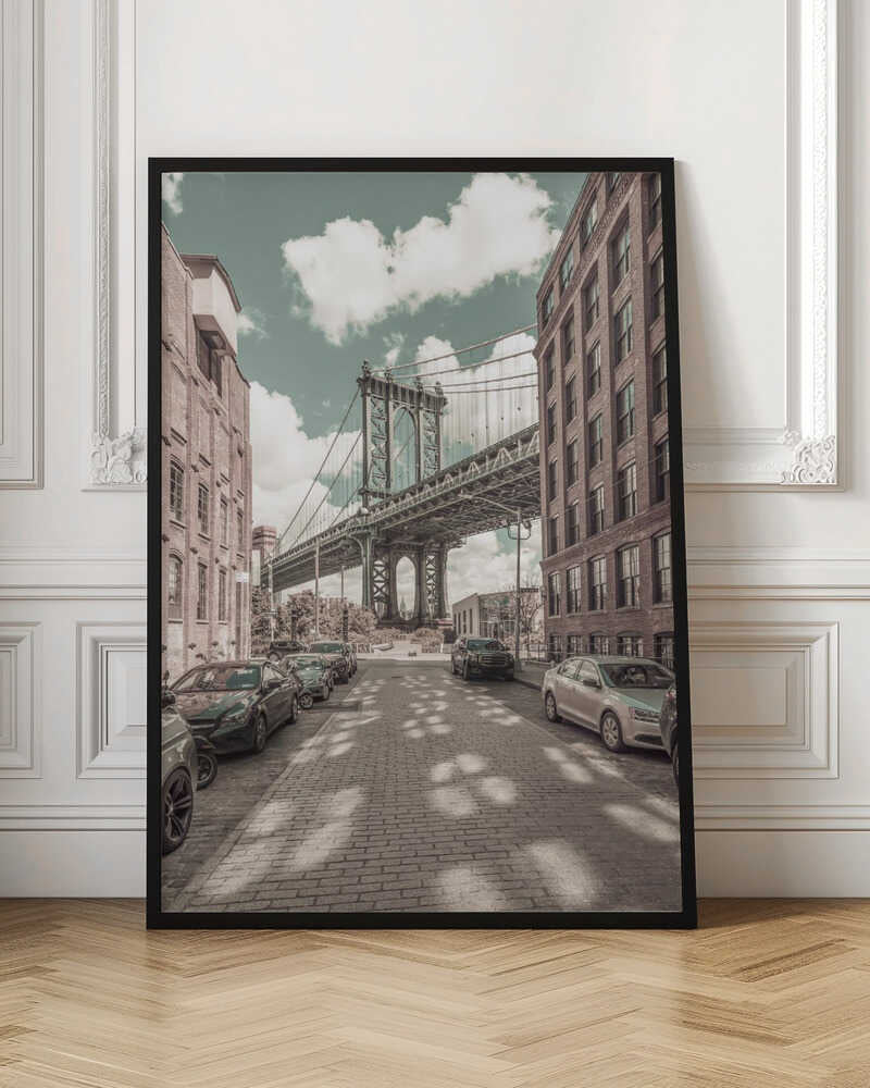 A vertical photograph of the Manhattan Bridge from a cobblestone street in DUMBO, Brooklyn, framed by tall brick buildings. The image has a vintage aesthetic with a muted teal sky and sepia-toned buildings and streets. Print