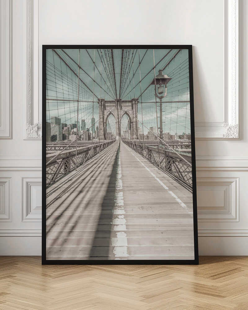 A vintage-style photograph looking down the center of the Brooklyn Bridge's wooden pedestrian walkway towards its iconic stone arches, with the New York City skyline visible in the background under a pale teal sky. The bridge's intricate network of suspension cables creates a strong sense of vanishing point perspective. Decor