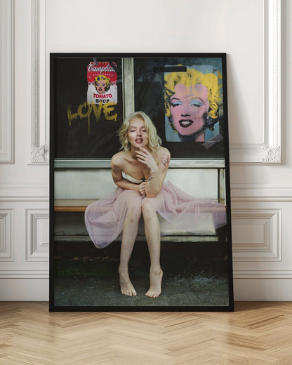 A blonde woman styled like Marilyn Monroe sits on a bench, wearing a pink tulle skirt. Behind her, a dark window features an Andy Warhol-style poster of Monroe, a Campbell's soup can parody, and the word 'LOVE' in gold graffiti. Artwork