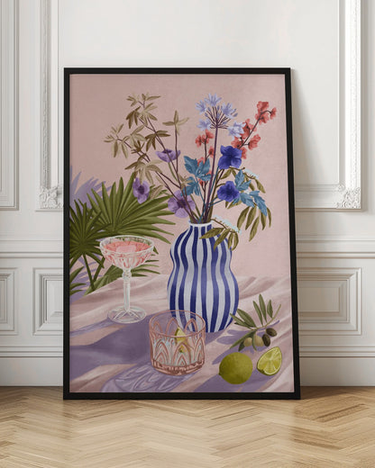A vibrant still life painting featuring a blue and white striped wavy vase filled with a colorful bouquet of flowers. To the left is a pink cocktail in a coupe glass and a decorative tumbler. In the foreground, there are fresh limes and an olive branch. The scene is set on a draped pink cloth against a soft pink background with shadows from palm leaves. Artwork