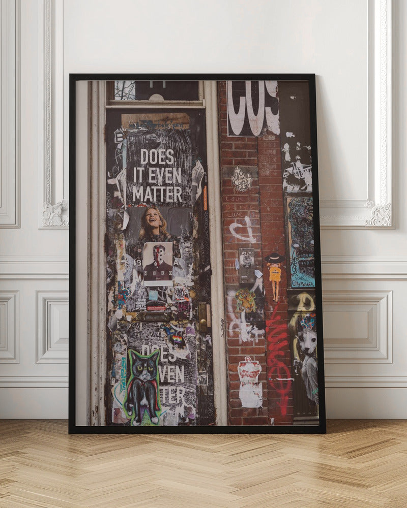 A close-up of a weathered dark door and an adjacent brick wall, both covered in a dense collage of street art, including peeling posters, stickers, and graffiti. A prominent poster on the door asks 'DOES IT EVEN MATTER' in bold white letters. Wall Art