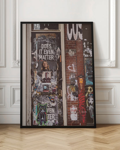 A close-up of a weathered dark door and an adjacent brick wall, both covered in a dense collage of street art, including peeling posters, stickers, and graffiti. A prominent poster on the door asks 'DOES IT EVEN MATTER' in bold white letters. Wall Art