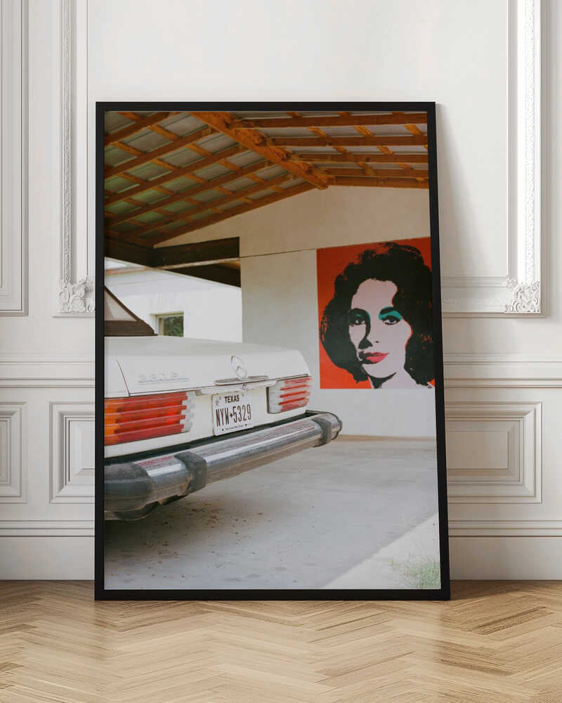 A film-style photograph showing the rear of a classic white Mercedes-Benz parked under a carport with exposed wooden beams. On the adjacent white wall hangs a vibrant red pop-art portrait of a woman with dark hair. Artwork