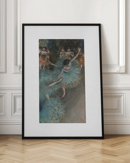 A framed art print of an impressionist painting depicting several ballet dancers on a stage. The central figure is a ballerina in a voluminous light blue tutu, viewed from a high angle. She is mid-pose with arms outstretched. Other dancers in orange and blue costumes are visible in the blurry background. Poster