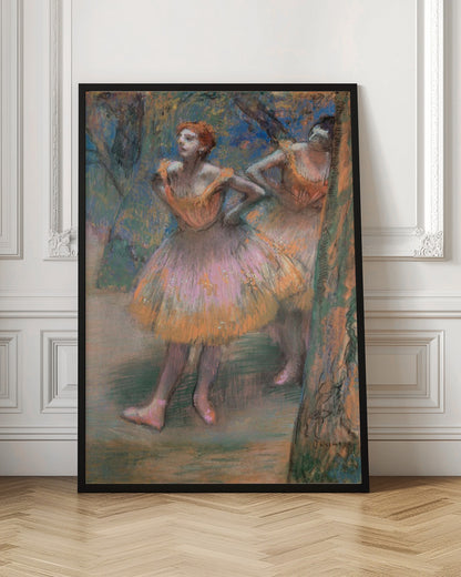 An impressionist pastel painting by Edgar Degas titled 'Dancers in Pink and Orange.' It depicts two ballerinas backstage. The dancer in the foreground has red hair, wears a pink and orange tutu, and stands with her hands on her hips, looking off to the side. A second dancer is partially visible behind her. The background is a textured blend of blues, greens, and browns. Artwork