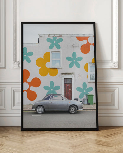 A retro-style gray convertible parked on a city street in front of a building with a large, colorful pop-art mural of stylized flowers in orange, yellow, and turquoise. Decor