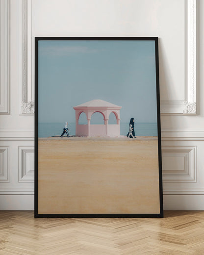 A minimalist photograph of a coastal scene with a pastel color palette. A pink arched gazebo sits on a promenade with the blue sea and sky in the background. A large expanse of beige sand is in the foreground. Two figures in hijabs walk in opposite directions on either side of the gazebo, one with a small child. Decor
