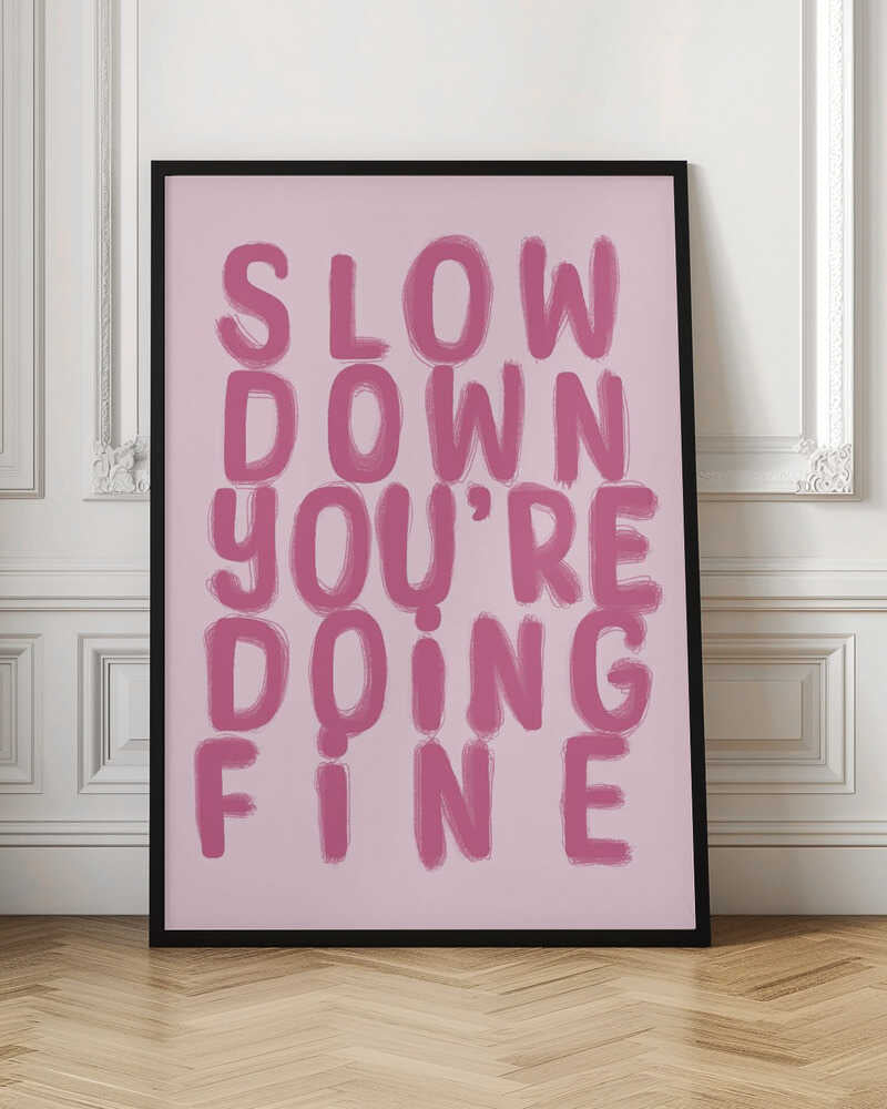 A framed art print featuring the phrase 'SLOW DOWN YOU'RE DOING FINE' in a bold, dark pink, brush-stroke font. The words are stacked vertically against a light pink background, all enclosed within a simple black frame. Artwork