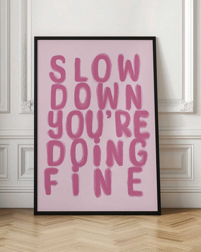 A framed art print featuring the phrase 'SLOW DOWN YOU'RE DOING FINE' in a bold, dark pink, brush-stroke font. The words are stacked vertically against a light pink background, all enclosed within a simple black frame. Artwork