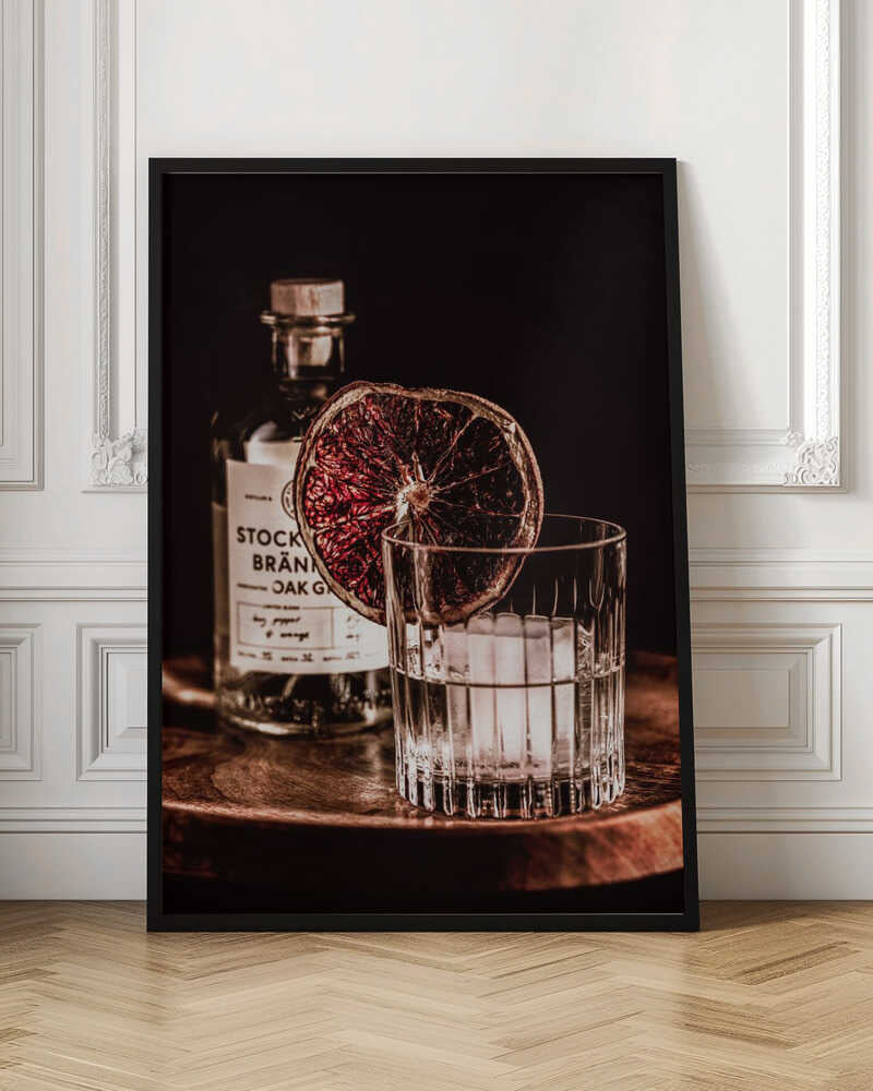 A moody still life photograph of a cocktail in a cut-crystal glass filled with ice. A dried blood orange slice garnishes the rim. In the soft-focus background, a bottle of gin rests on a dark wooden tray against a black backdrop. Wall Art