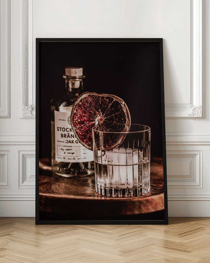 A moody still life photograph of a cocktail in a cut-crystal glass filled with ice. A dried blood orange slice garnishes the rim. In the soft-focus background, a bottle of gin rests on a dark wooden tray against a black backdrop. Wall Art