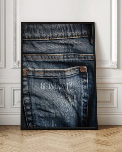 A detailed close-up photograph of a faded blue jean back pocket, showing the texture of the denim, orange stitching, and a copper rivet. Faint white text is stenciled onto the pocket. Poster