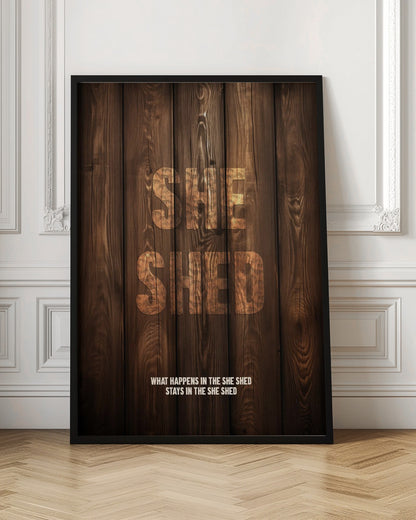 A rustic poster with a dark brown wood panel background. The words 'SHE SHED' are in large, blocky letters with a distressed wood texture. Below, a smaller caption reads 'WHAT HAPPENS IN THE SHE SHED STAYS IN THE SHE SHED'. The poster is displayed in a thin black frame. Print