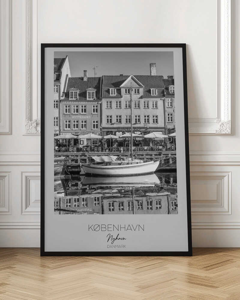 A black and white framed poster of Nyhavn in Copenhagen. The photograph features classic Danish buildings lining a canal, with a sailboat moored in the water. The buildings and boat are perfectly reflected in the still water. At the bottom, text reads 'KØBENHAVN', 'Nyhavn', and 'DANMARK'. Wall Art