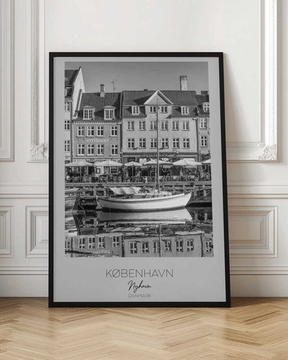 A black and white framed poster of Nyhavn in Copenhagen. The photograph features classic Danish buildings lining a canal, with a sailboat moored in the water. The buildings and boat are perfectly reflected in the still water. At the bottom, text reads 'KØBENHAVN', 'Nyhavn', and 'DANMARK'. Wall Art