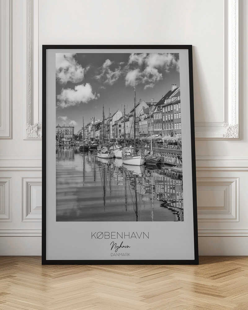 A black and white poster of Nyhavn harbor in Copenhagen. The photograph shows traditional sailboats and ships moored along a canal, with a line of historic, multi-story buildings on the far bank. The sky has fluffy clouds, and the entire scene is reflected in the calm water. The poster has a white border with the text 'KØBENHAVN', 'Nyhavn', and 'DANMARK' at the bottom, and is shown within a black frame. Poster