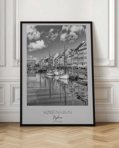 A black and white poster of Nyhavn harbor in Copenhagen. The photograph shows traditional sailboats and ships moored along a canal, with a line of historic, multi-story buildings on the far bank. The sky has fluffy clouds, and the entire scene is reflected in the calm water. The poster has a white border with the text 'KØBENHAVN', 'Nyhavn', and 'DANMARK' at the bottom, and is shown within a black frame. Poster