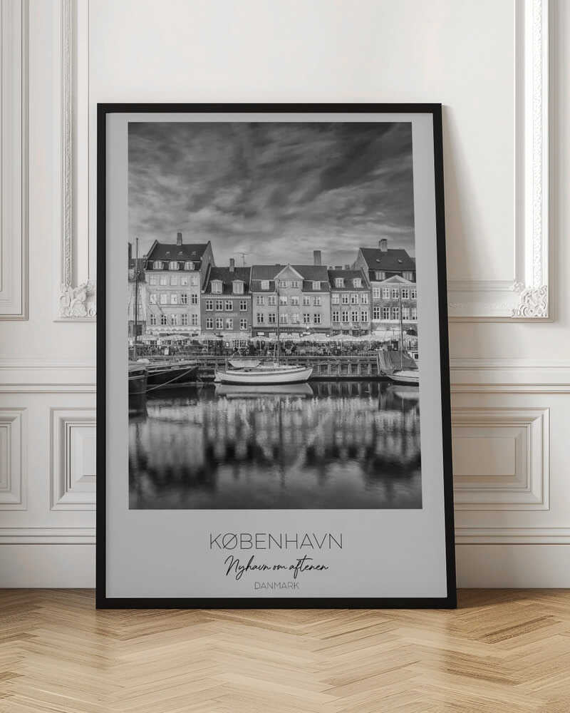 A framed black and white photograph of the Nyhavn harbor in Copenhagen, Denmark. The image shows a row of classic gabled houses along the waterfront, with several sailboats moored in the canal. The buildings and a dramatic cloudy sky are reflected in the water. At the bottom, a white border contains the text 'KØBENHAVN', 'Nyhavn om aftenen', and 'DANMARK'. Wall Art