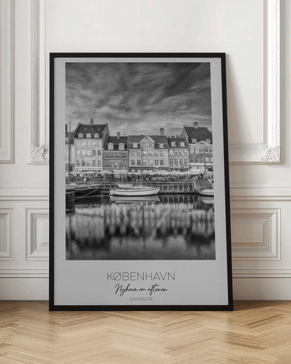 A framed black and white photograph of the Nyhavn harbor in Copenhagen, Denmark. The image shows a row of classic gabled houses along the waterfront, with several sailboats moored in the canal. The buildings and a dramatic cloudy sky are reflected in the water. At the bottom, a white border contains the text 'KØBENHAVN', 'Nyhavn om aftenen', and 'DANMARK'. Wall Art