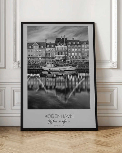 A black and white poster of Nyhavn harbor in Copenhagen at night. Historic buildings with glowing windows line the canal, and their lights create shimmering reflections in the water. A boat is docked in the foreground. The poster has text at the bottom that reads 'KØBENHAVN, Nyhavn om aftenen, DANMARK'. Poster