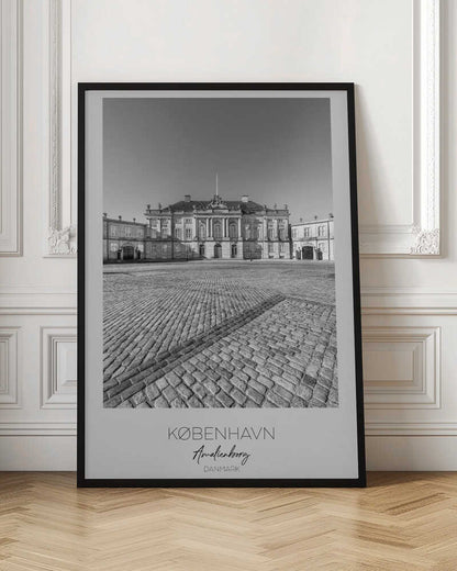 A framed, vertical black and white photograph of Amalienborg Palace in Copenhagen. The shot is taken from a low angle, emphasizing the large cobblestone square in the foreground. The palace stands symmetrically in the background under a clear sky. At the bottom of the poster, text reads 'KØBENHAVN', 'Amalienborg', and 'DANMARK'. Poster