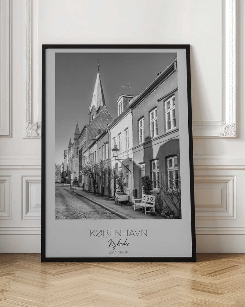 A black and white framed travel poster of a charming cobblestone street in Nyboder, Copenhagen. The photograph shows a sunlit row of historic houses with a prominent church steeple in the background. At the bottom of the poster, the words 'KØBENHAVN', 'Nyboder', and 'DANMARK' are written. Wall Art