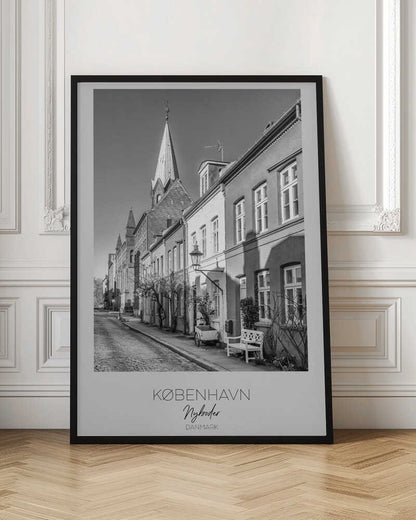 A black and white framed travel poster of a charming cobblestone street in Nyboder, Copenhagen. The photograph shows a sunlit row of historic houses with a prominent church steeple in the background. At the bottom of the poster, the words 'KØBENHAVN', 'Nyboder', and 'DANMARK' are written. Wall Art