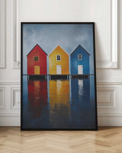 A painting of three houses in primary colors—red, yellow, and blue—lined up on a pier over a body of water. The houses and their vibrant colors are reflected in the calm water below, all under a muted, overcast sky. The piece is in a black frame. Decor