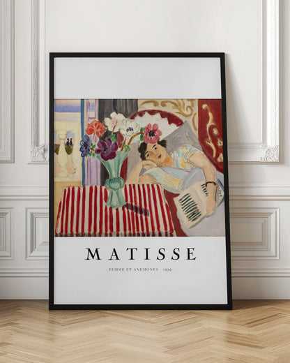 A poster featuring Henri Matisse's 1939 painting, 'Femme et Anemones'. The artwork depicts a woman in a colorful top reclining on a couch, holding a book. Beside her, on a red and white striped tablecloth, sits a vase filled with vibrant anemone flowers. The poster has a black frame and white matting, with 'MATISSE' printed below the image. Artwork