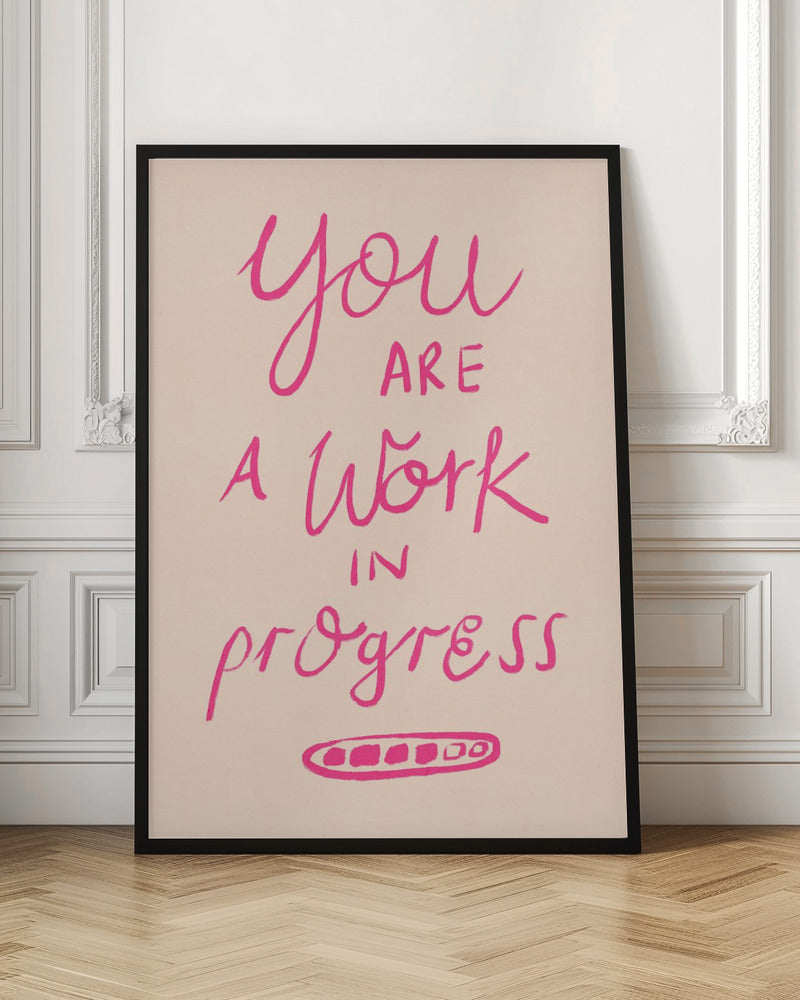 A framed art print featuring the motivational quote 'You are a work in progress'. The text is rendered in a vibrant pink, hand-lettered style against a soft beige background. The words 'You', 'a Work', and 'progress' are in a flowing script, while 'are' and 'in' are in a simple uppercase font. Wall Art