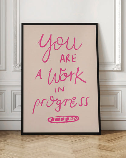 A framed art print featuring the motivational quote 'You are a work in progress'. The text is rendered in a vibrant pink, hand-lettered style against a soft beige background. The words 'You', 'a Work', and 'progress' are in a flowing script, while 'are' and 'in' are in a simple uppercase font. Wall Art