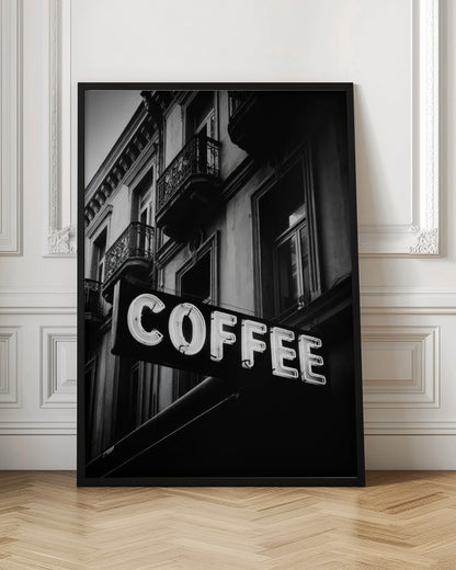 A dramatic, low-angle, black and white photograph of a bright white neon sign that reads 'COFFEE', mounted on a classic-style building with ornate balconies. Print
