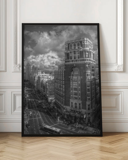 A dramatic black and white high-angle photograph of a bustling city street. A large, ornate historical building dominates the right side, while traffic flows along the avenue on the left under a sky filled with heavy, textured clouds. Poster