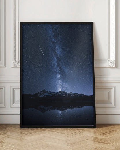 A breathtaking night photograph of the Milky Way galaxy stretching vertically across a deep blue, star-filled sky. Two shooting stars streak through the atmosphere above a dark, snow-dusted mountain range, which is perfectly reflected in the calm, mirror-like surface of a lake in the foreground. Artwork