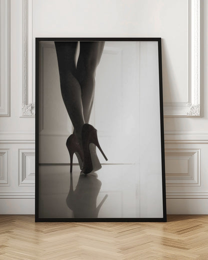 A low-angle, black and white photograph focusing on a woman's legs as she stands with them crossed. She wears dark red stiletto high heels, which cast a clear reflection on the glossy floor. Her legs are silhouetted against a bright white door in the background, creating a dramatic and sensual mood. Decor