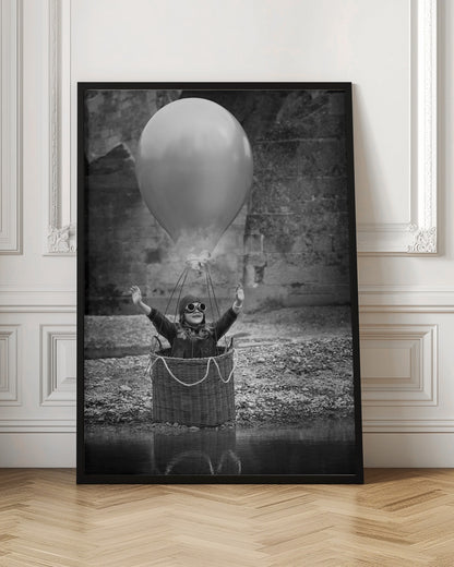 A whimsical black and white photograph of a young girl wearing an aviator cap and goggles, joyfully raising her arms in the air while standing in a wicker basket attached to a large balloon, mimicking a hot air balloon. The scene is set by the water with a stone wall in the background. Poster