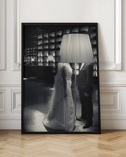 A surreal black and white photograph of a bride and groom whose heads are comically obscured by a large, pleated lampshade from a floor lamp standing between them. They are in a modern, dimly lit room with a wall of illuminated shelves. Poster