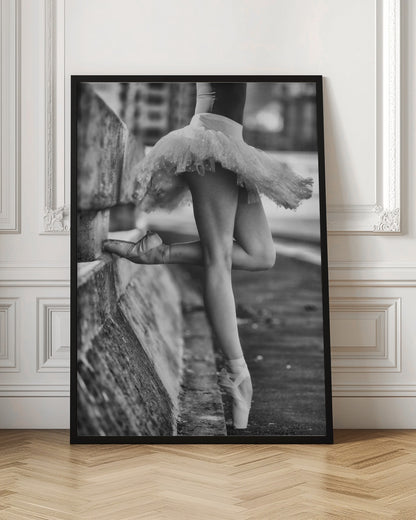 A black and white photograph focusing on the lower half of a ballerina in a tutu and pointe shoes. She stands en pointe with one foot on the ground, while the other foot rests against a rough, concrete curb, highlighting the contrast between delicate dance and an urban environment. Decor