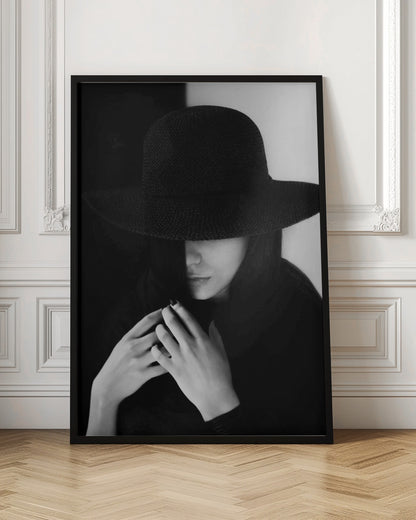 A mysterious black and white portrait of a woman wearing a large, black, wide-brimmed hat that casts a deep shadow over her eyes. Her hands are clasped at her chest, and the background is split vertically into black and white sections. Artwork