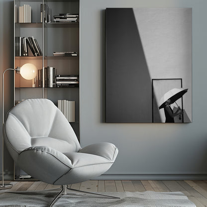 A minimalist black and white photograph of a woman in a black dress holding a two-toned umbrella. She stands in front of a door against a light wall, with a sharp, diagonal shadow dramatically cutting across the upper left of the image, creating a high-contrast, geometric composition. Decor